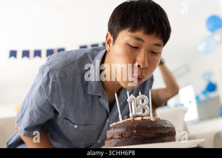 Asiatischer Mann, der Kerzen auf der Geburtstagskuche bläst und zu Hause den 18. Geburtstag feiert Stockfoto