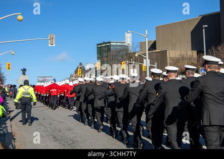 Dieses Foto zeigt Militärpersonal, einschließlich Marinesoldaten und Soldaten in roter Uniform, die in einer Parade entlang einer Stadtstraße unter klarem blauem Himmel marschieren. Stockfoto