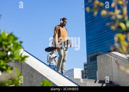 afroamerikanischer Mann, der auf den plaza Steps geht, Skateboard trägt, Rucksack mit Kopfhörern Stockfoto