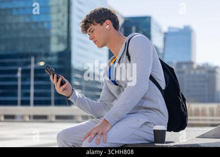 Mann mit Hoodie, der auf der plaza sitzt und Smartphone, Ohrstöpsel, Rucksack, Kaffeetasse hält Stockfoto