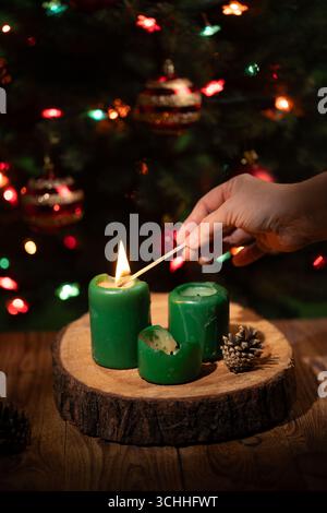 Eine Hand zündet mit einem Streichholz grüne Kerzen an, die auf einer Holzplatte vor einem Weihnachtsbaum mit funkelnden Lichtern und Ornamenten angeordnet sind. Stockfoto
