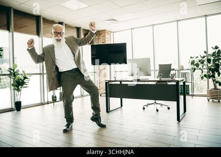 Reifer Geschäftsmann in eleganter Kleidung tanzt fröhlich in einem modernen Büro mit Schreibtisch und Computer Stockfoto