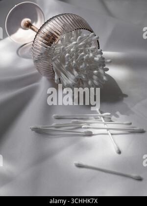 Wattestäbchen aus Ohr, die aus einem transparenten Glas auf eine weiße Tischdecke gelangen. Stockfoto