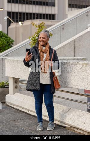 Ältere afroamerikanerin, die ihr Smartphone auf der Terrasse hält, mit einer braunen Ledertasche Stockfoto