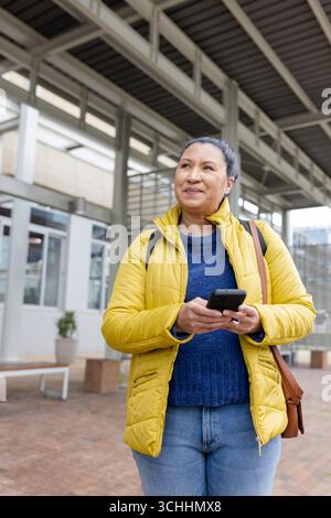Frau in gelber Jacke auf dem Gehweg unter dem Metalldach, die das Smartphone überprüft, Ledertasche in der Hand hält Stockfoto
