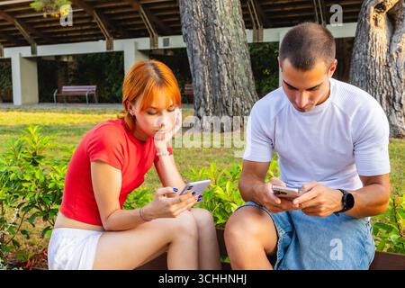 Junge Paare, die sich gegenseitig ignorieren, während sie Smartphones im Park benutzen Stockfoto