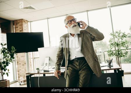 Ein älterer Geschäftsmann in eleganter Jacke, der einen Anruf führt, während er sich an einem Arbeitsplatz auf seinen Schreibtisch lehnt Stockfoto