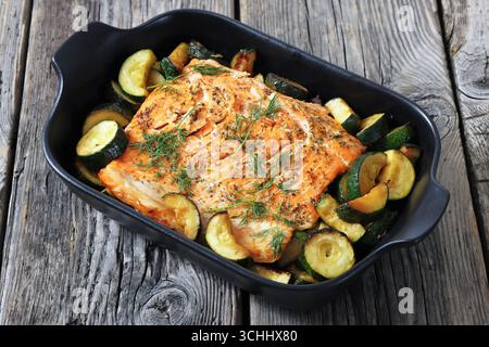 Gebacken im Ofen Lachsfilet mit Zucchini und Dill in schwarzer Auflaufform auf rustikalem Holztisch, horizontale Ansicht, Nahaufnahme, nicht KI generiert Stockfoto