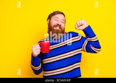 Fröhlicher Mann in gestreiftem Pullover, der einen roten Becher vor einem leuchtend gelben Hintergrund hält und fröhliche Energie ausstrahlt Stockfoto
