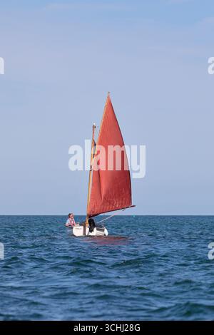 Ein kleines grün-weißes Segelboot namens „Catcher“ mit roten Segeln und einem Außenbordmotor, der auf dem offenen Meer segelt Stockfoto