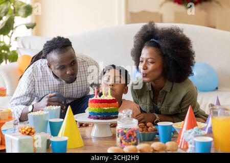 Familie feiert zu Hause Geburtstag und bläst zusammen Kerzen auf buntem Kuchen aus Stockfoto