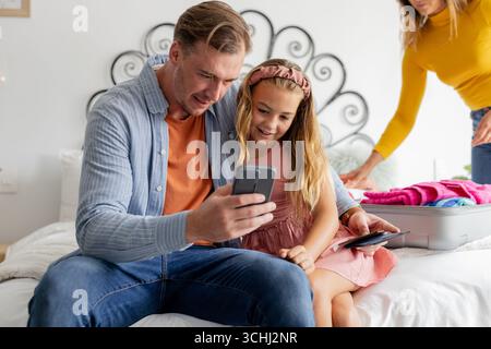 Vater und Tochter benutzen das Smartphone im Bett, während Mutter Koffer in der Nähe packt Stockfoto