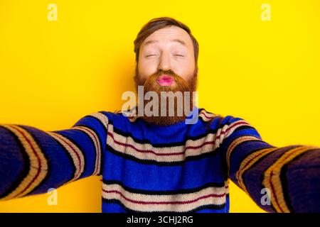 Fröhlicher junger Mann mit rotem Bart posiert glücklich auf hellgelbem Hintergrund, trägt einen gestreiften Pullover und macht eine lustige Geste Stockfoto