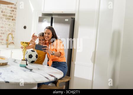 Mutter und Tochter machen Selfie mit Fußball-Trophäe in der Küche und lächeln glücklich Stockfoto