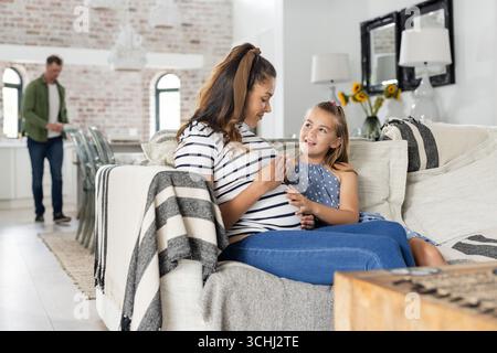 Schwangere Mutter und Tochter sitzen auf der Couch, lächeln und knüpfen zu Hause Stockfoto