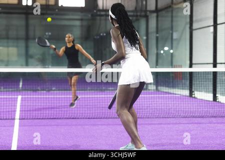 Verschiedene weibliche Gegner, die sich in der Sporthalle über das Netz stellen, spielen auf dem Padel-Court den Ball Stockfoto