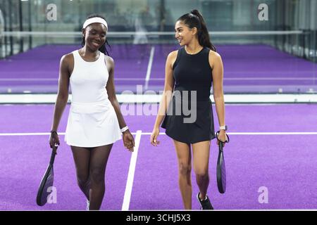Verschiedene Freundinnen in Sportbekleidung, die über den violetten Paddle-Tennisplatz laufen und Schläger halten Stockfoto