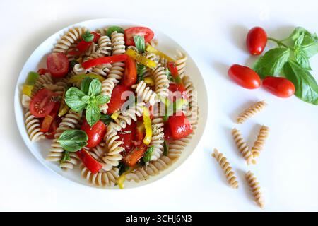 Vollkornnudelsalat mit Paprika, Tomaten, Olivenöl, Oregano und frischen Basilikumblättern auf weißem Hintergrund. Gesund und vegetarisch. Sommergericht. Stockfoto