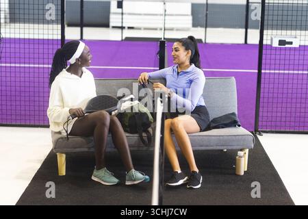 Verschiedene Freundinnen sitzen auf einer Bank auf dem lila Platz mit Pickleball-Paddeln in der Nähe von Sporttaschen Stockfoto