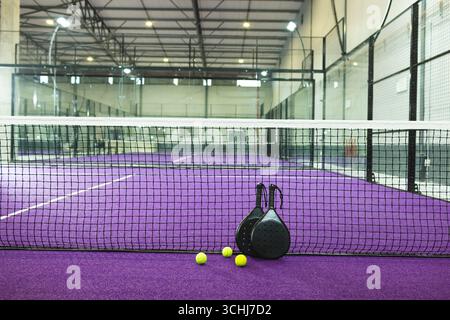 Zwei schwarze Padel-Schläger lehnen sich mit dem Netz auf violetten Rasen, während drei gelbe Kugeln liegen Stockfoto