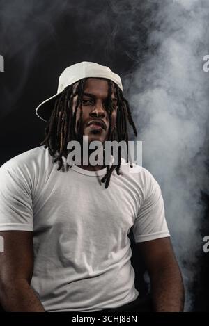 Mann mit Dreadlocks, der Hut und weißes Hemd trägt, posiert für die Kamera in einem Studio vor einem rauchigen Hintergrund. Selbstbewusster und ernster junger Mann. Stockfoto