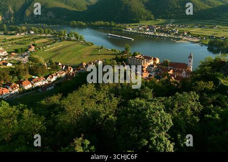 Sonniger Hochwinkelblick auf Dürnstein mit roten Häusern, Weinbergen, Hügeln, Donau mit vorbeifahrendem Binnenschiff und Campingplatz gegenüber Stockfoto