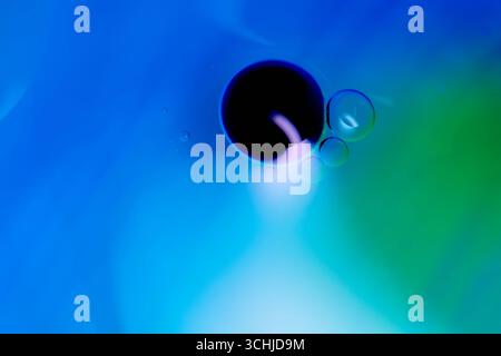 Schwarzer Öltropfen mit drei kleinen Blasen auf einem blau-grünen Verlaufshintergrund, Makroaufnahme mit Oberflächenspannung und glatten Farbübergängen. Stockfoto