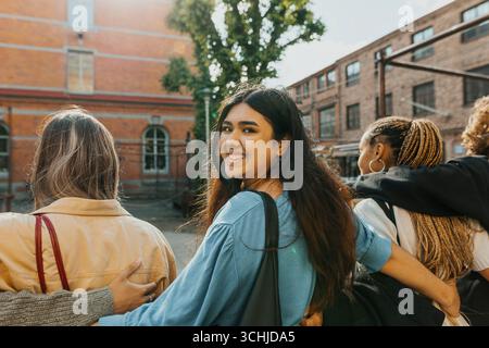 Porträt einer lächelnden jungen Frau, die mit dem Arm um Freunde geht, während sie über die Schulter schaut Stockfoto