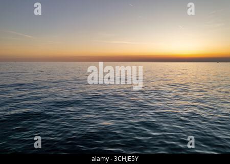 Eine ruhige, endlose Meereslandschaft, die den goldenen Sonnenuntergang über dem ruhigen Meereswasser einfängt. Sanfte Wellen reflektieren warme Orange- und Gelbtöne am friedlichen Horizont Stockfoto
