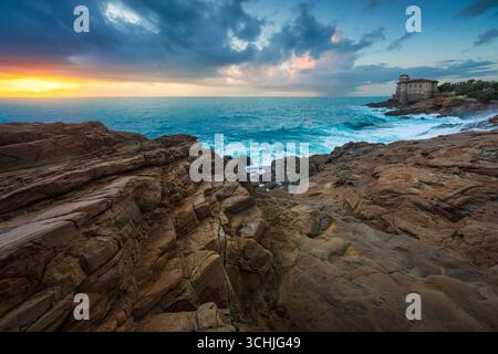 Das historische Schloss Boccale thront an der dramatischen felsigen Küste in der Nähe von Livorno während des stürmischen Sonnenuntergangs mit Wellen gegen Klippen. Toskana, Ital Stockfoto