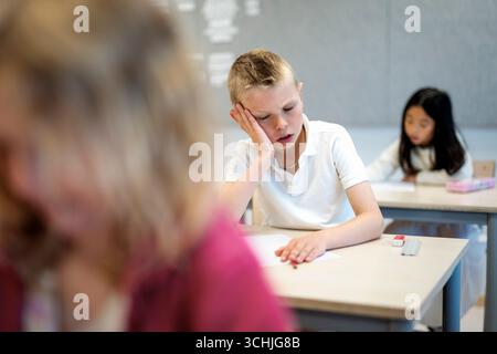 Müder Junge, der sich auf den Ellenbogen gestützt hat, während er im Grundschulzimmer am Schreibtisch saß Stockfoto