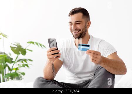 Lächelnder junger mann aus europa, der im weißen T-Shirt online einkaufen kann, bestellt Waren auf dem Smartphone und zeigt seine Kreditkarte Stockfoto