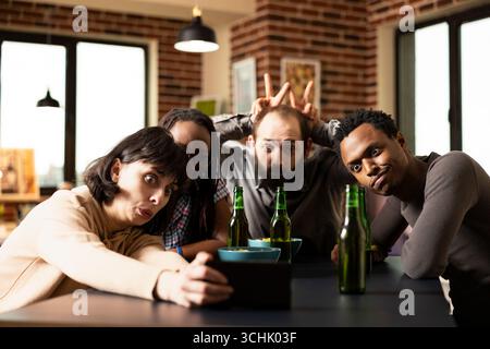 Verschiedene Freunde machen beim Selfies am Tisch einen blöden Gesichtsausdruck, während der bärtige Mann spielerisch Hasenohren zeigt. Multiethnische Gruppen sitzen zusammen, posieren mit dem Smartphone und schaffen lustige Erinnerungen. Stockfoto