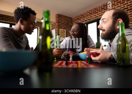 afroamerikanische Individuen und kaukasier, die sich während des Brettspiels unterhalten. Multiethnische Gruppe von Freunden, die sich mit Snacks und Getränken um den Tisch herum zusammensetzten und sich miteinander verbinden. Stockfoto