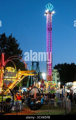 Amherst, MA - USA - 23. August 2025 Eine lebendige Karnevalsszene in der Abenddämmerung mit einem riesigen Drop Ride, einem hell beleuchteten Riesenrad und anderen leuchtenden Attrac Stockfoto