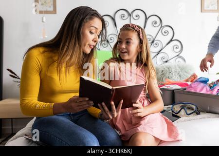 Mutter liest Buch mit Tochter im Bett, beide lächeln und genießen die gemeinsame Zeit Stockfoto