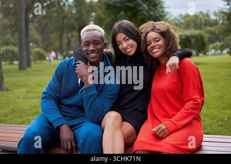 Freunde im Teenageralter mit mehreren ethnischen Gruppen. afroamerikanisch-asiatischer Student verbringt Zeit miteinander Multirassische Freundschaft glücklich lächelnde Menschen gekleidet bunte Sportbekleidung Sitzbank Park im Freien Stockfoto