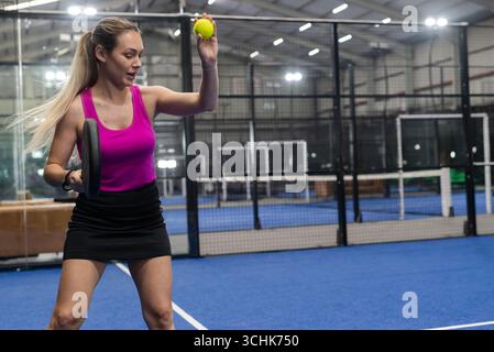 Weiße Frau, die Tennisball hochhebt, während sie auf dem Padel-Platz hinter Zäunen steht Stockfoto