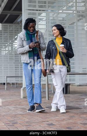Verschiedene Freunde laufen Seite an Seite durch den urbanen plaza mit Smartphone, Kaffeetasse, Skateboard Stockfoto