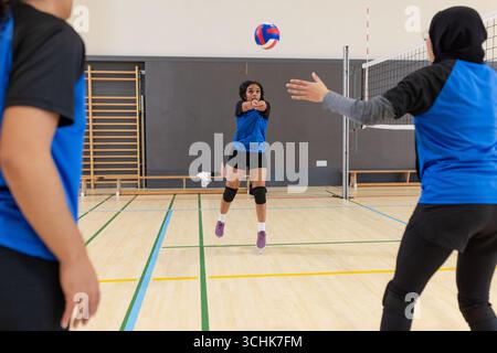 Vielfältiges weibliches Team in blauen Trikots, das Volleyball über das Netz im Fitnessstudio mit Wandstangen schlägt Stockfoto