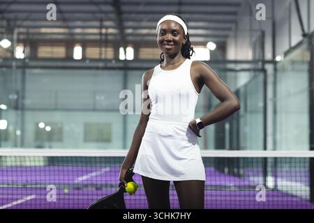 afroamerikanische Tennisspielerin, die auf einem violetten Platz neben dem Netz steht und Schläger und Ball hält Stockfoto