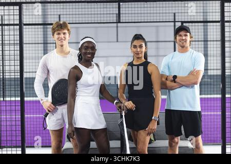 Verschiedene Padel-Teamkollegen stehen am Netz auf dem violetten Platz mit Paddeln mit Bank und Tasche dahinter Stockfoto