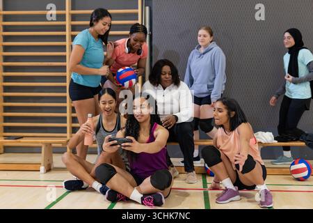 Verschiedene weibliche Volleyball-Mannschaftskameraden treffen sich auf der Bank im Fitnessstudio mit Volleyball und Smartphone Stockfoto