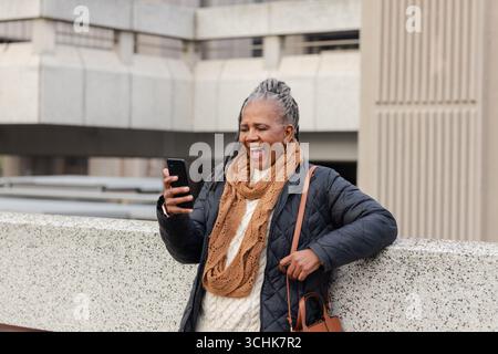 Ältere afroamerikanerin, die Jacke trägt, sich auf das Geländer stützt, Telefon hält, braune Tasche Stockfoto