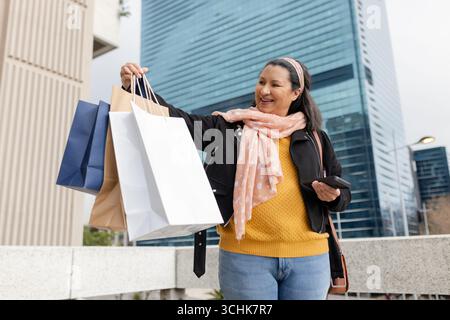 Frau mit Papiertaschen, die ihr Smartphone auf dem Stadtrand mit Wolkenkratzern überprüft Stockfoto