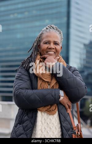 Ältere afroamerikanerin lächelt und steht auf dem stadtplatz in Jacke, mit Smartwatch Stockfoto