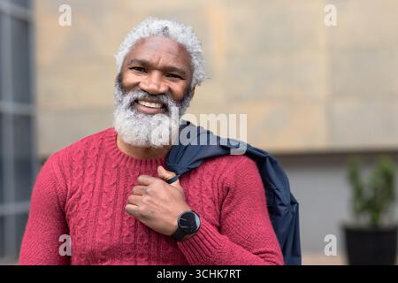 Ranghoher afroamerikanischer Mann, der vor einer Steinmauer steht und einen roten Pullover trägt, der eine Jacke hält Stockfoto