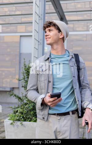 Mann, der auf dem Bürgersteig der Stadt vor dem modernen Gebäude steht und ein Smartphone hält und den Koffer zieht Stockfoto