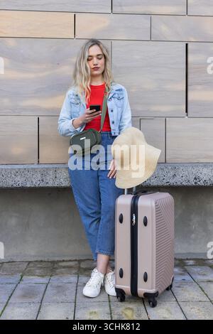 Frau mit Smartphone, die auf dem Bürgersteig neben dem Koffer steht und mit Strohsonnenhut steht Stockfoto