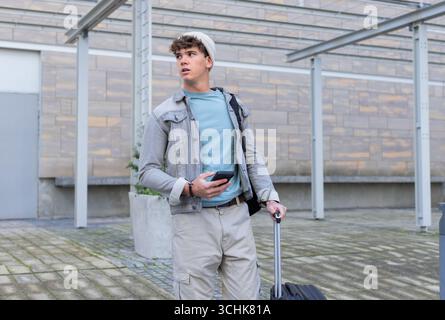 Mann, der Smartphone hält und den Koffer packt, Schultertasche unter dem Dach am Transitknoten trägt Stockfoto